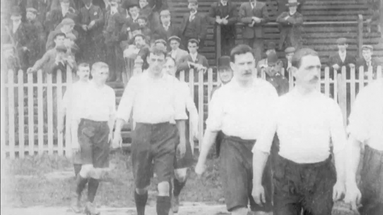 An Edwardian Bolton Wanderers football team file onto a threadbare pitch.