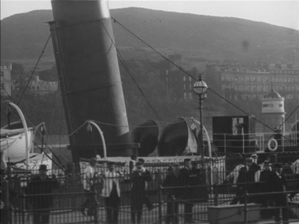 Welcoming crowds greet the arrival of a steamer to the Isle of Man, seen from a stunning perspective on deck.