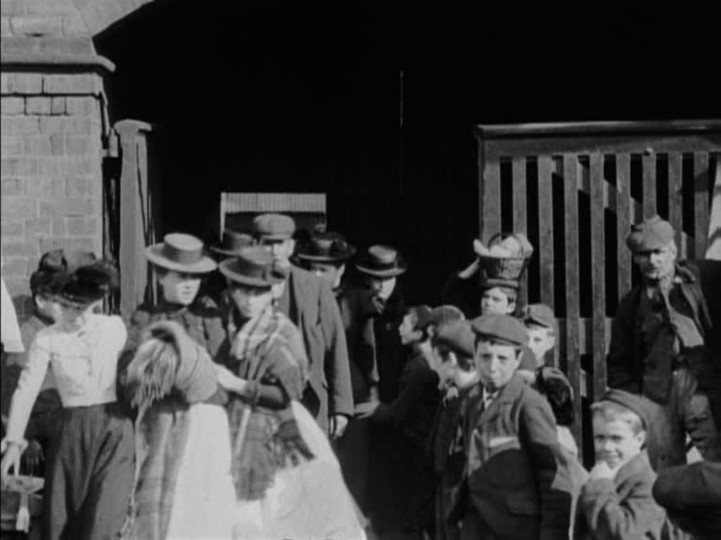 Smartly-dressed men and women leave an unknown (textiles?) factory at lunchtime.